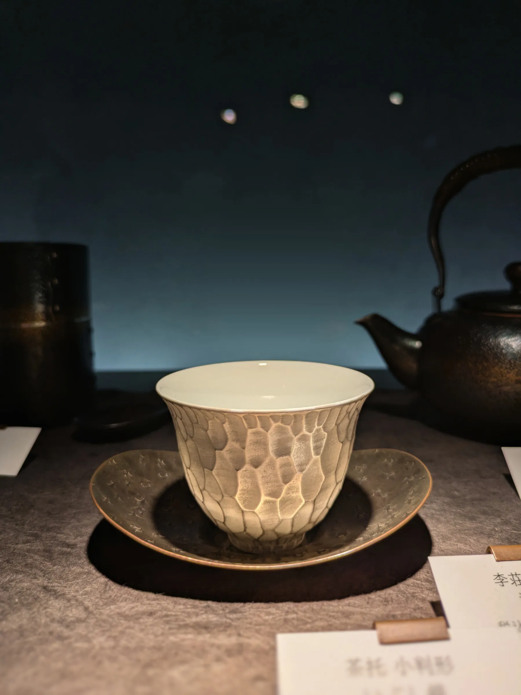 展示台上的白色杯子與茶托，杯身外側是銀色的塊面紋理，後方有茶罐與鐵壺。