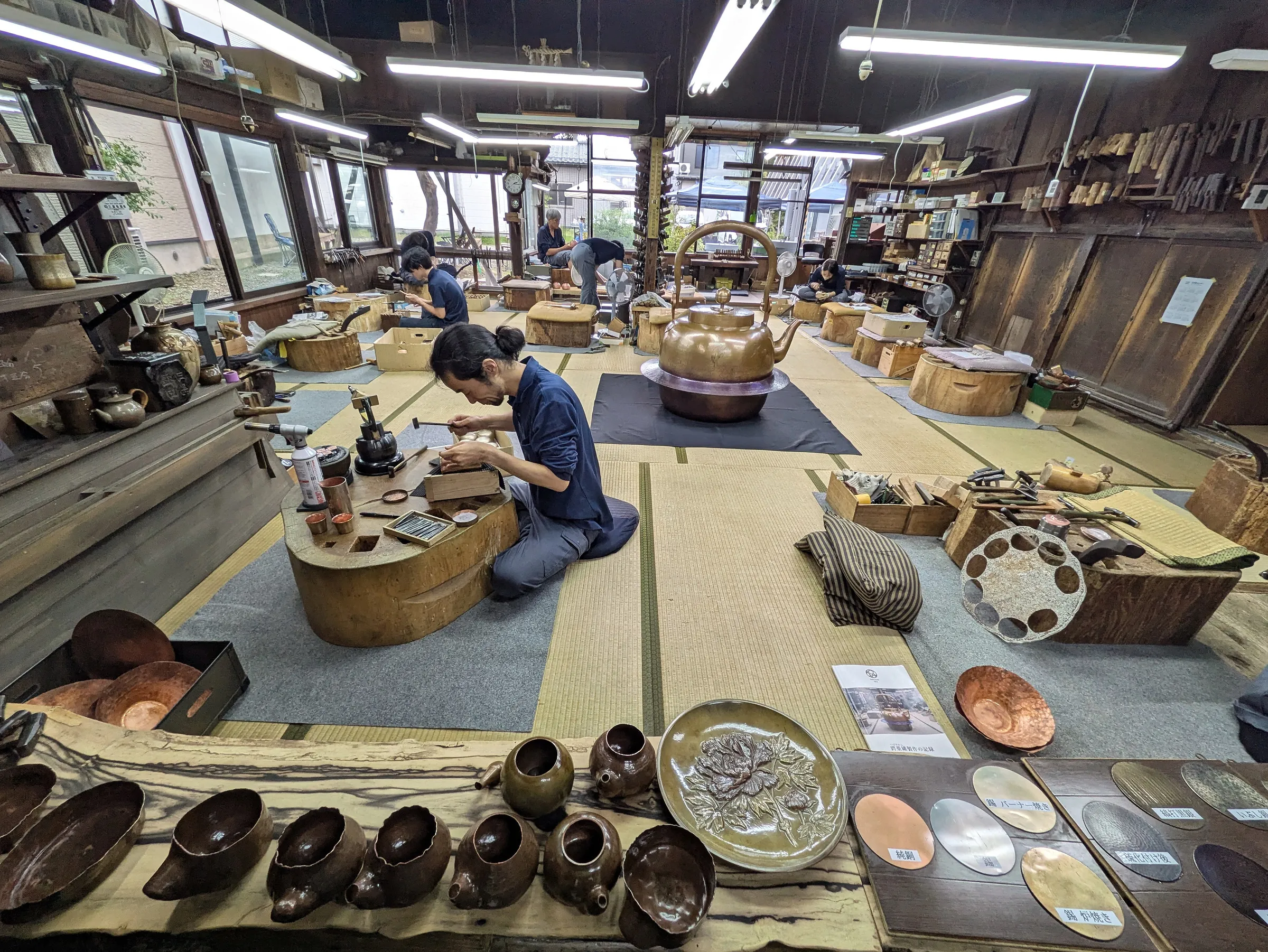 玉川堂工房全景：職人坐在榻榻米上敲打銅器，前方長桌排著製作中的器物與金屬板樣品。